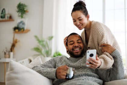 Jong stel zit ontspannen op de bank en bekijkt samen iets op een smartphone, passend bij het volgen van hypotheekrentes of financiële updates.